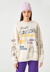 Sudadera color crema con impresiones gráficas en varios colores, con un corte oversize, puños acanalados y múltiples diseños de texto juguetones.