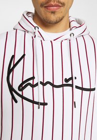 Felpa con cappuccio bianca con strisce verticali rosse e nere, con un grande logo nero ricamato. Include coulisse bianche e dettagli metallici.