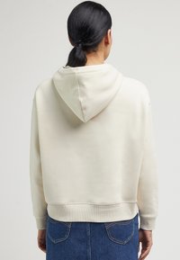 Felpa crema realizzata in morbido tessuto, con un ampio cappuccio, polsini e orlo a coste. Indossata con una gonna di jeans blu, che mostra una vestibilità morbida.