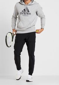 Sudadera gris con el logotipo negro "adidas Tennis", combinada con pantalones deportivos negros y zapatillas blancas. Sosteniendo una raqueta de tenis y una pelota.