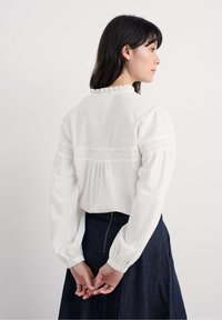 Frau mit dunklen Haaren, die eine weiße Bluse mit langen Ärmeln und Spitzenverzierungen sowie einen dunklen Jeansrock trägt, steht mit hinter dem Rücken verschränkten Händen.