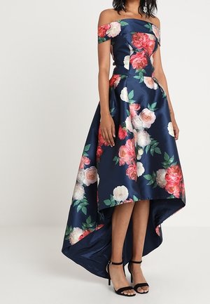 Marineblå satin kjole med blomsterprint i pink og hvid, med off-shoulder design, høj-lav kant og sorte ankelremssandaler.
