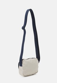 Borsa tracolla in tessuto beige con tracolla regolabile blu navy con il marchio "TOMMY JEANS", chiusura con cerniera e forma morbida e arrotondata.
