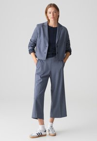 Blazer gris cintré avec un bouton unique, associé à un pantalon large gris. Des baskets blanches avec des rayures bleues et des chaussettes texturées complètent le look.