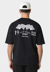 Fekete rövid ujjú póló, fehér grafikákkal: versenyt flaggok, "19 PAS DE MONACO 88" és "Atelier Racing" felirat. Klasszikus pamut anyag.