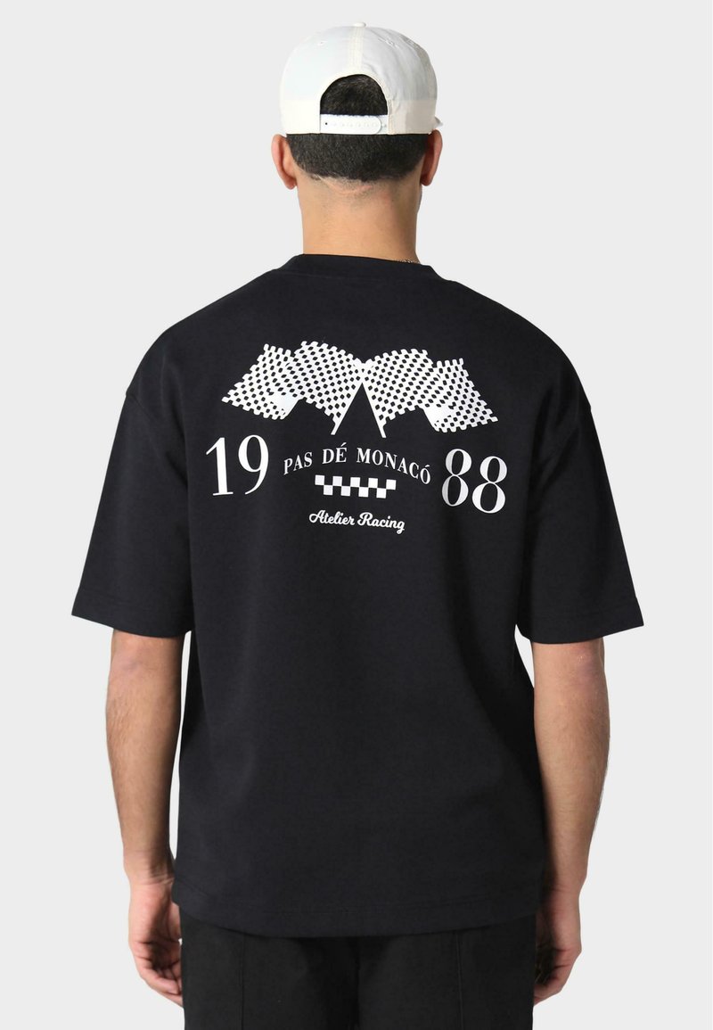 Fekete rövid ujjú póló, fehér grafikákkal: versenyt flaggok, "19 PAS DE MONACO 88" és "Atelier Racing" felirat. Klasszikus pamut anyag.