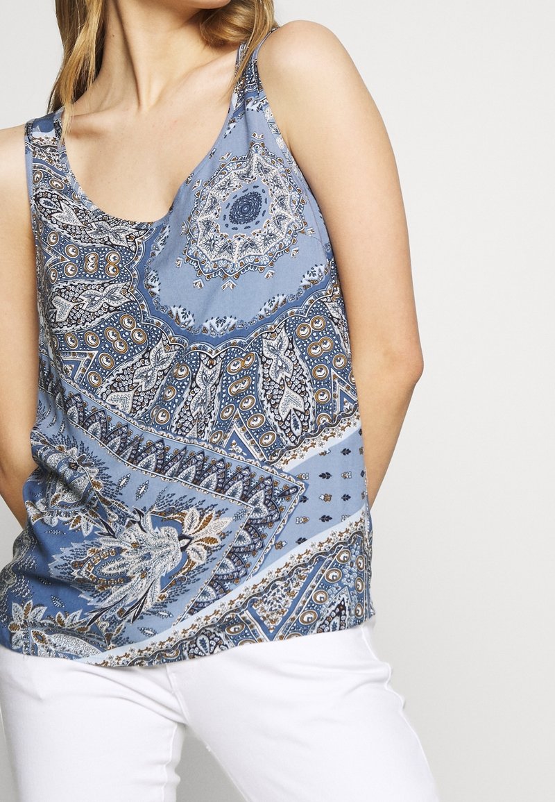 Femme portant un top bleu sans manches avec un motif paisley détaillé blanc, noir et or, associé à un pantalon blanc sur un fond clair.
