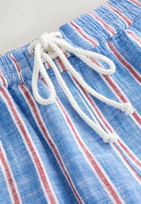 Primer plano de pantalones cortos de tela con rayas azules, rojas y blancas, con cordón trenzado blanco atado en un lazo sobre una cintura elástica.