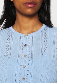 Cárdigan de punto azul claro con un cuello redondo, que presenta un cierre de botones, patrón de encaje y textura acanalada.