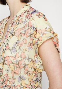 Gros plan sur une personne portant un chemisier transparent à manches courtes avec un motif de papillons multicolores dans des teintes pastel.