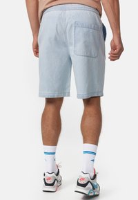 Pantalones cortos de mezclilla de color azul claro con cintura elástica y bolsillo trasero. Presentan dobladillos enrollados y un corte relajado. Llevados con zapatillas blancas y grises.