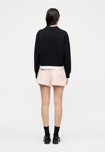 Γυναίκα που στέκεται με την πλάτη γυρισμένη, φορώντας μαύρο μακρυμάνικο τοπ, λευκό εσώρουχο, ροζ υφασμένα σορτς, μαύρες κάλτσες και μαύρα παπούτσια σε λευκό φόντο.