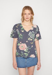 Blusa de manga curta com padrão floral em tecido azul escuro, apresentando grandes flores pink e verdes, decote em V e um corte solto, combinada com uma saia de ganga.