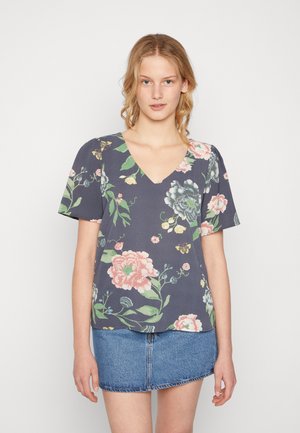Blusa de manga curta com padrão floral em tecido azul escuro, apresentando grandes flores pink e verdes, decote em V e um corte solto, combinada com uma saia de ganga.