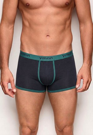 Torso masculino con calzoncillos tipo bóxer negros con cintura y ribete verdes, de pie frente a un fondo claro y liso.