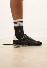 Sort atletisk sko lavet af ruskind og syntetisk materiale, med en cremefarvet hæl, parret med sort-hvide stribede strømper.