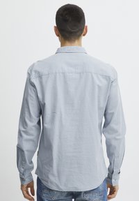 Lys blå langermet skjorte, myk stoff, uformell krage og knappede mansjetter. Rundet nederkant med et merke på venstre side. Brukt med jeans.