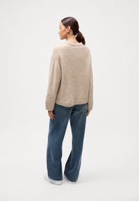Béžový vlnený sveter s uvoľneným strihom, okrúhlym výstrihom a rebrovaným lemom, v kombinácii s voľnými modrými džínsami a bielymi teniskami.