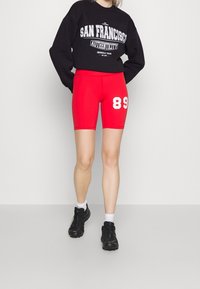 Pantalones cortos rojos de bicicleta con una cintura alta, que presentan un gran "89" blanco en el lateral. Combinados con una sudadera negra y zapatillas negras.