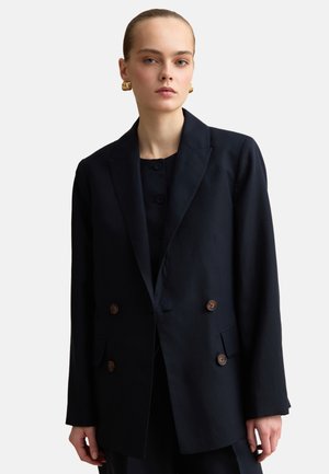 Jeune femme aux cheveux lissés en arrière portant des boucles d'oreilles en anneau dorées et un blazer noir croisé avec des boutons marron.