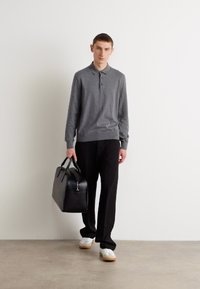Polo a maniche lunghe grigio, pantaloni neri, tiene una borsa in pelle nera e sneakers bianche. Design semplice e aderente con colletto classico.