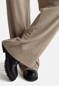 Béžové vlněné kalhoty širokého střihu s žebrovanými manžetami, v kombinaci s černými koženými botami s kulatou špičkou a texturovanou podrážkou pro lepší přilnavost.