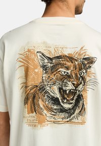 Camiseta de color crema que presenta un gráfico de un gato agresivo en negro y naranja, con un fondo texturizado y un texto manuscrito debajo.