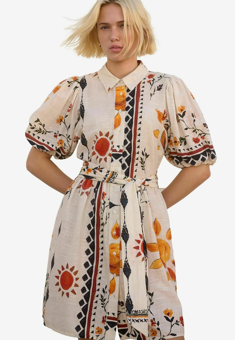 Kurzes Blumenkleid aus beigem Stoff, mit Puffärmel, Kragen, durchgehender Knopfleiste und einem Gürtel, verziert mit bunten Sonnen- und Blumenmustern.