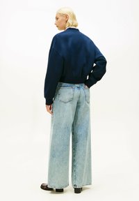 Mujer con cabello rubio corto, lleva un suéter corto azul marino, jeans de pierna ancha de color azul claro y zapatos de tacón marrón oscuro, de pie contra un fondo blanco.