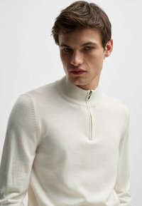 Light cream zip-up sweater made of a smooth, fine-knit material. Features a high collar and subtle stitching detailing along the shoulders.