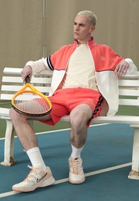 Sportski set koji se sastoji od koralno-crvene i bijele jakne s patentnim zatvaračem i kontrastnim prugama, u kombinaciji s koralnim kratkim hlačama. Drži žuti tenis reket.