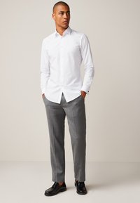 Hvid skjorte med lange ærmer og spids krave, kombineret med skræddersyede grå bukser og sorte loafers. Enkel, ren design med knapper.