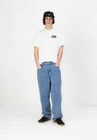 Hvid kortærmet T-shirt med grafik, blå løstsiddende jeans og sorte sneakers. Sort kasket tilføjer kontrast til ensemblet.