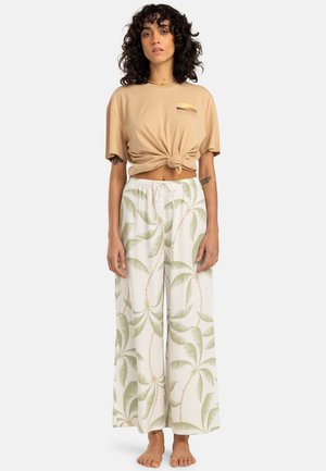 Beige knotted t-shirt with a logo, paired with white wide-leg pants featuring green palm leaf patterns and soft, lightweight fabric.