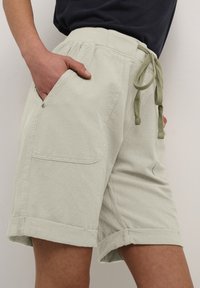 Hellgrüne Shorts aus strukturiertem Stoff. Sie verfügen über einen elastischen Bund, einen Kordelzug und umgeschlagene Säume. Eine seitliche Taschen-Detail ist sichtbar.