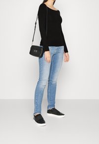 Frau trägt ein schwarzes Langarmoberteil, hellblaue Skinny Jeans, schwarze Slip-On-Sneaker und hält eine schwarze Calvin Klein Umhängetasche.