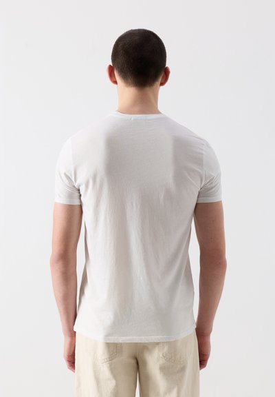 Homme aux cheveux courts et foncés, de dos, portant un T-shirt blanc uni et un pantalon beige clair, sur un fond blanc uni.
