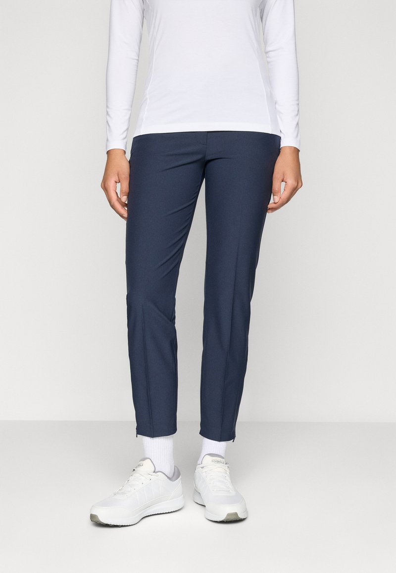 Persoană îmbrăcată cu pantaloni slim bleumarin, bluză lungă albă, șosete albe și adidași albi, stând în fața unui fundal simplu.
