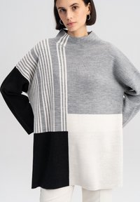 Grijze en zwarte oversized trui met een hoge hals, voorzien van verticale witte strepen en contrasterende vlakken in verschillende grijstinten en zwart.