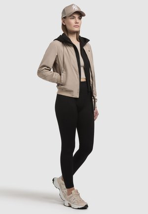 Kvinde iført beige kasket, beige jakke med sort inderlag, sorte leggings og beige sneakers, stående med den ene hånd i jakkelommen.