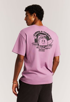 Lila katoenen T-shirt met een grafische print op de achterkant van een cartoon taco en surfplank, tegen een contrasterende zwarte achtergrond.