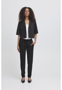 Svart cropped blazer med en enkelt knapp, sammen med svarte bukser med snøring og flate, firkantede svarte sko. Strukturert hår.