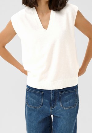 Femme portant un haut en maille blanc sans manches avec un col en V et un jean bleu, les mains posées sur les hanches devant un fond uni.