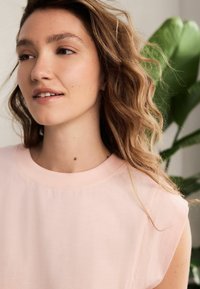 Světle růžový bezrukávový top s širokým kulatým výstřihem, vyrobený z měkké látky. Přirozený make-up, vlnité vlasy; zelená rostlina v pozadí.