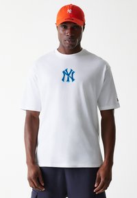 T-shirt en coton blanc avec un logo bleu et noir des New York Yankees sur la poitrine, porté avec une casquette orange. Manches courtes, coupe décontractée.