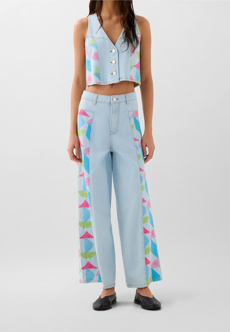 Veste en jean bleu clair raccourcie avec boutons sur le devant, associée à un pantalon en jean à jambes larges avec des motifs géométriques colorés sur les côtés.