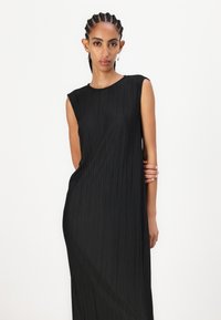 Rochie neagră, plisată, cu decolteu rotund și design fără mâneci. Materialul are un aspect texturat, cu linii verticale.