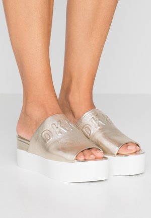 Metalen gouden slip-on sandalen met plateauzool en open tenen, voorzien van een gegraveerd "DKNY" logo op een glad bovenwerk en een dikke witte zool.