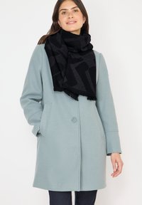 Femme portant un manteau bleu clair à un bouton et une écharpe noire à motifs, se tenant avec une main dans la poche devant un fond blanc.