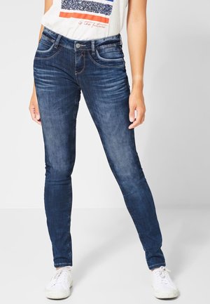 Frau trägt dunkelblaue Skinny-Jeans mit Ausbleichungen und weiße Sneakers, steht mit gespreizten Beinen vor einem schlichten Hintergrund.